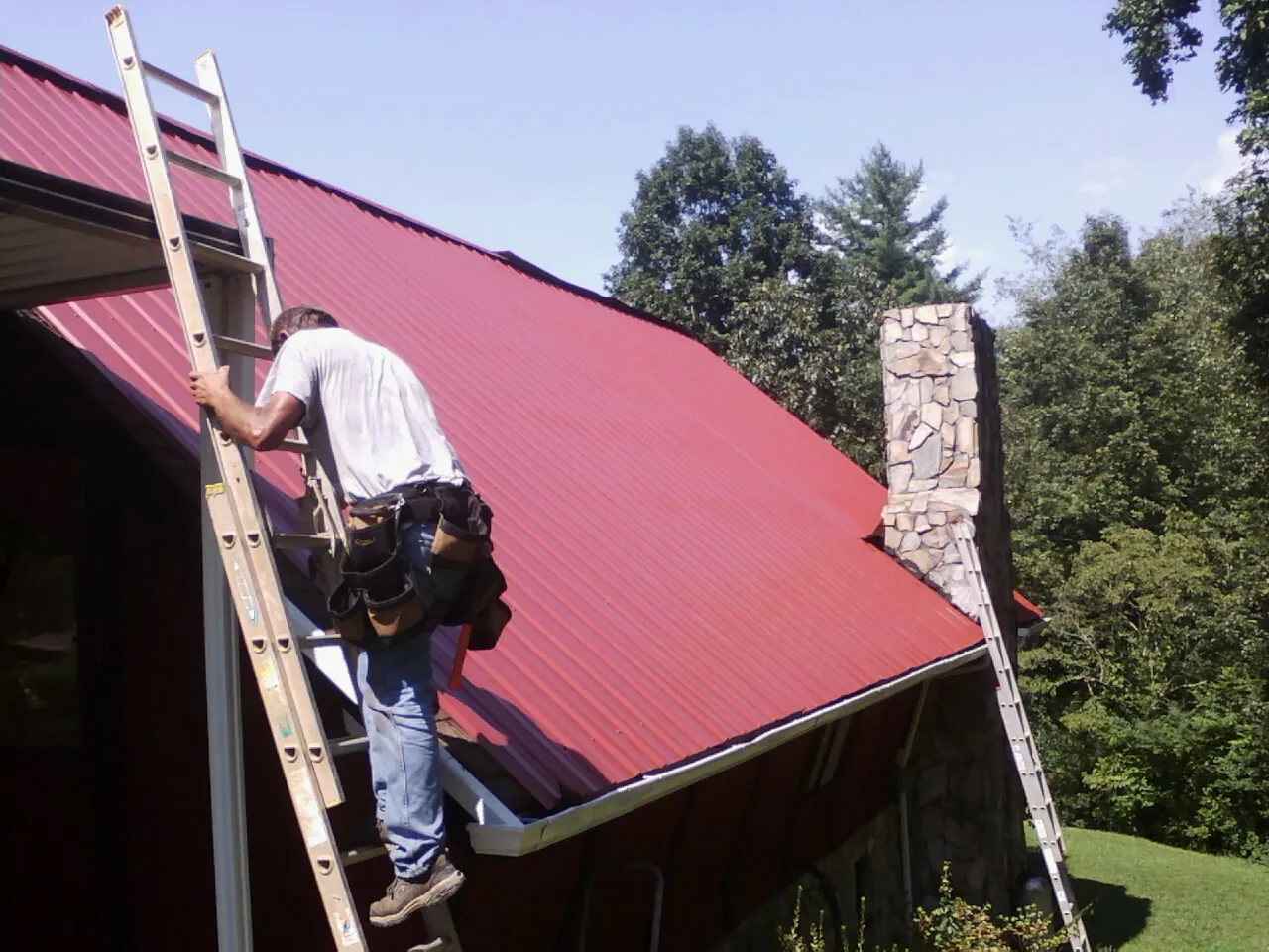 Roofer on a ladder beside a red metal roof and stone chimney — Soffit & Fascia Repair by Roofing Brothers in Croghan